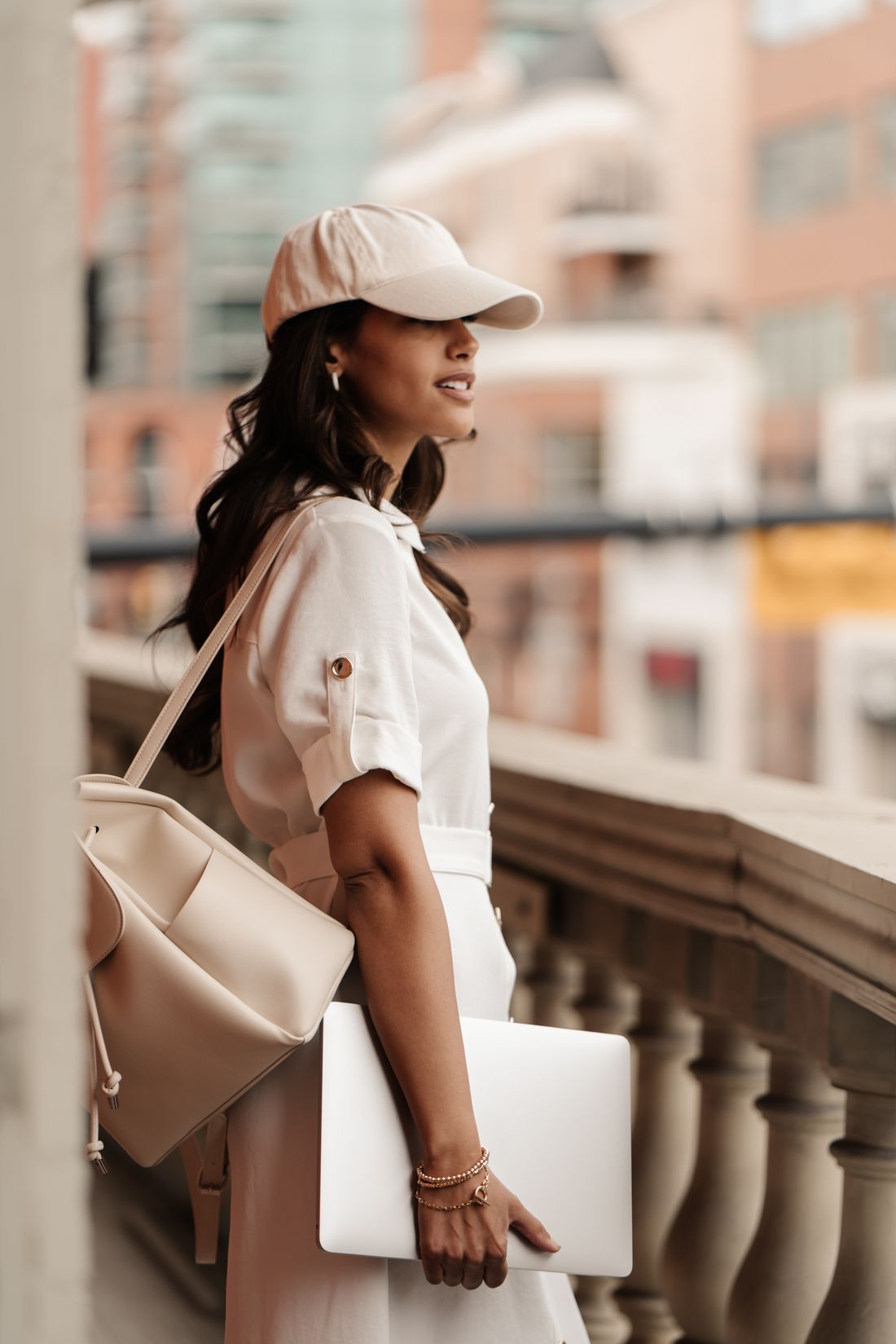 A woman in a cap and white shirt on a balcony above a city street, laptop under her arm, looking ahead.