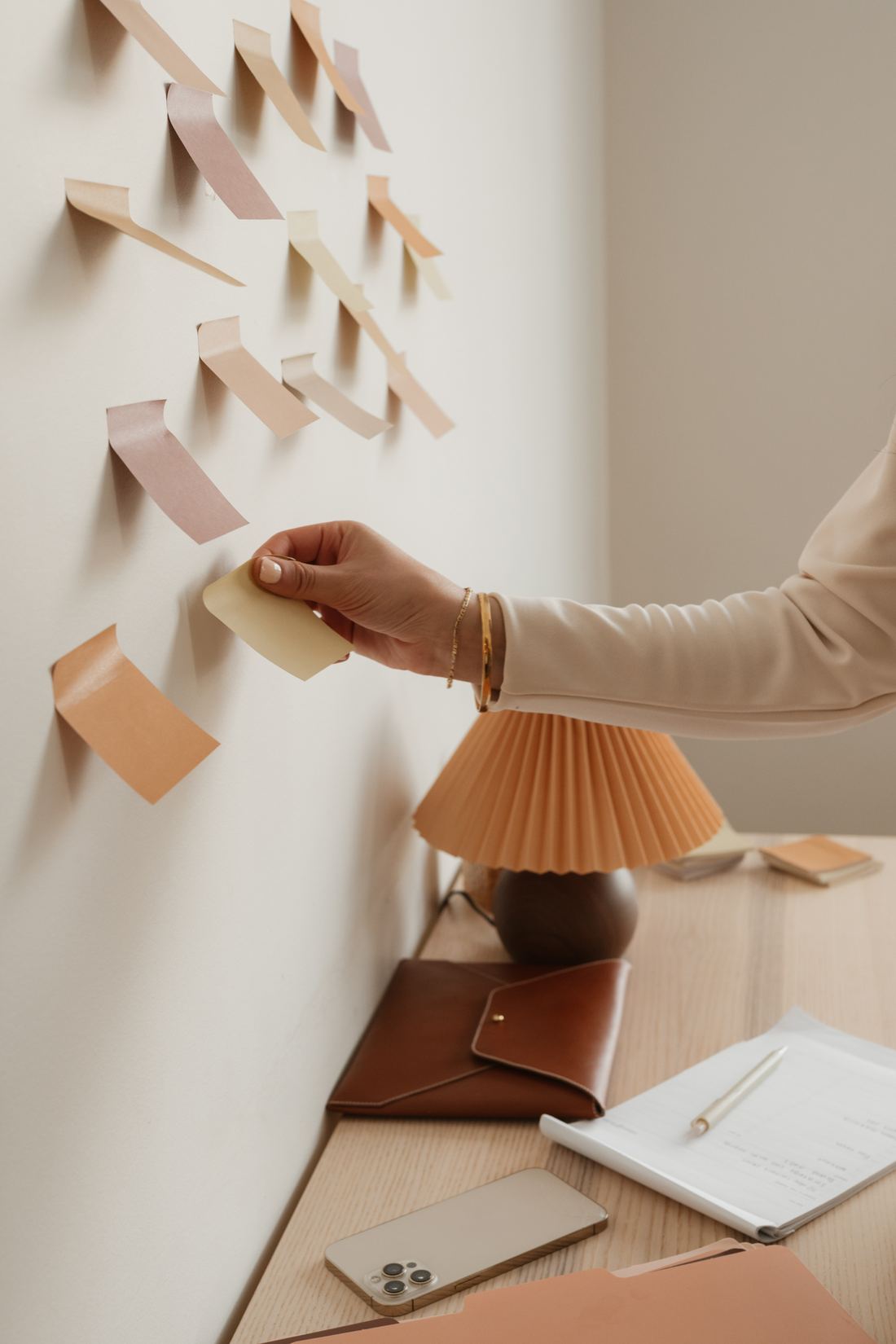 A hand placing a blank note on a wall of loosely arranged paper notes above a desk.